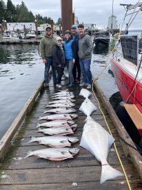 A group of five anglers proudly displaying their catch of multiple Salmon and a Halibut on the dock after a successful fishing trip.