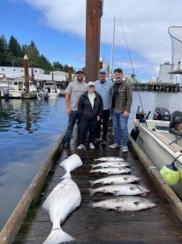 A group of four anglers proudly displaying their catch of the day, featuring a large Halibut and several Salmon, at a dock in a scenic fishing environment.