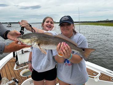 Two young anglers proudly displaying a large Redfish while fishing from a boat in a scenic coastal environment.