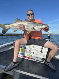 Angler proudly displaying a large Striped Bass while seated on a fishing boat, showcasing a sunny day on the water.