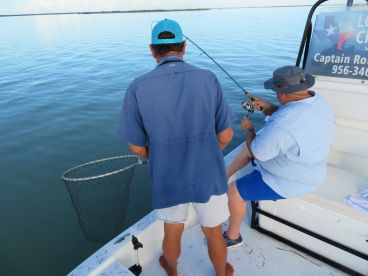 Two anglers engaged in fishing on a boat, with one preparing to net a catch while the other reels in a line, showcasing a serene fishing environment.