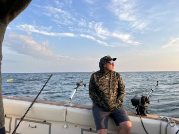 A man in a camouflage hoodie enjoying the view from the interior of a fishing boat, with fishing gear visible and a calm sea in the background.