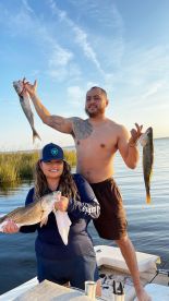 Two anglers proudly displaying their catches, a large Speckled Trout and a Redfish, while enjoying a fishing trip in a scenic coastal environment.