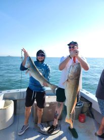 Two anglers proudly displaying their catches, a large Redfish and a Striped Bass, aboard a fishing charter in clear waters.
