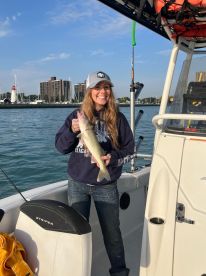 A smiling angler proudly displays a Walleye while fishing from a boat in a scenic harbor setting.