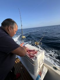 A fisherman filleting a freshly caught fish on a cooler aboard a fishing boat, with a clear view of the ocean in the background.