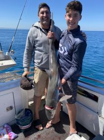 A young angler and an adult proudly displaying a large shark on a fishing boat, showcasing a successful day on the water.