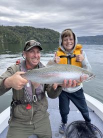 A young angler and an adult proudly display a large Rainbow Trout while fishing on a serene lake, showcasing a successful day on the water.