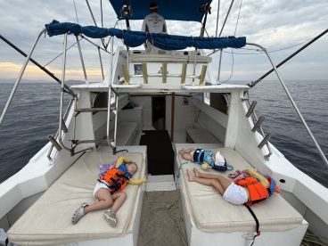 Two young children resting on the cushioned seating area of a fishing boat, wearing life jackets, while a crew member is visible in the background.