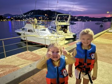 Two young girls in life jackets posing happily at the dock during twilight, with boats in the background.