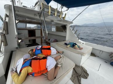 Two young children resting on the seating area of a fishing boat, wearing life jackets, while enjoying a day at sea.