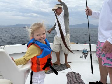 A young girl in a life jacket smiles while fishing on a boat, as an adult reels in a catch in the background, showcasing a fun family fishing experience.