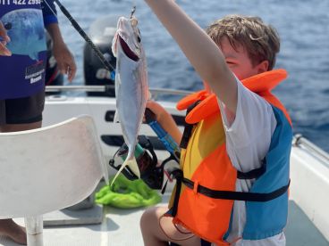 A young angler in a life jacket proudly displaying a caught fish while fishing on a boat in open waters.