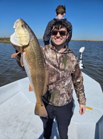 Angler proudly displaying a large Redfish while fishing on a calm day in a scenic waterway.