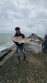 A young angler proudly displaying a large Redfish while fishing off a pier, with a cloudy sky and other anglers in the background.