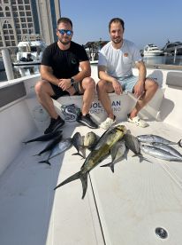 Two anglers sitting on a boat with a variety of fish, including a large Mahi Mahi, after a successful fishing trip in a marina setting.