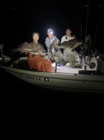 Three anglers proudly displaying their large Grouper catches while fishing at night on a boat.