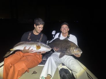 Two anglers proudly displaying their catches, a large Redfish and a Black Drum, while fishing at night.