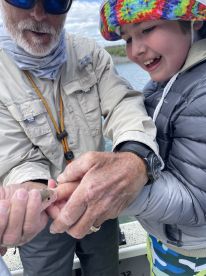 A young angler excitedly holds a small fish with the help of an adult, showcasing a memorable fishing moment on the water.