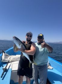 Two anglers proudly displaying a large Yellowtail while fishing on a boat in clear blue waters.