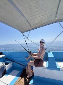 A female angler enjoying a fishing experience on a blue boat, actively reeling in a catch under a sunny sky.