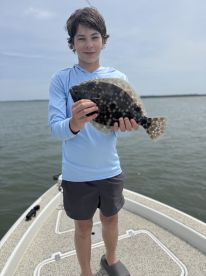 A young angler proudly displaying a Flounder while standing on the bow of a boat during a fishing trip.