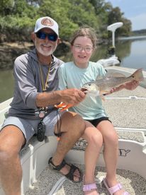 A young girl proudly holds a Redfish while fishing with an adult on a boat, showcasing a successful catch in a scenic waterway.