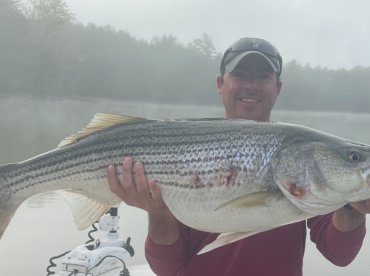 An angler proudly displaying a large striped bass while fishing on a misty lake.