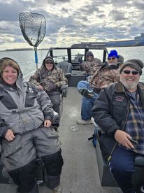 A group of anglers enjoying a day on a fishing boat, dressed for cooler weather, with a scenic backdrop of water and clouds.